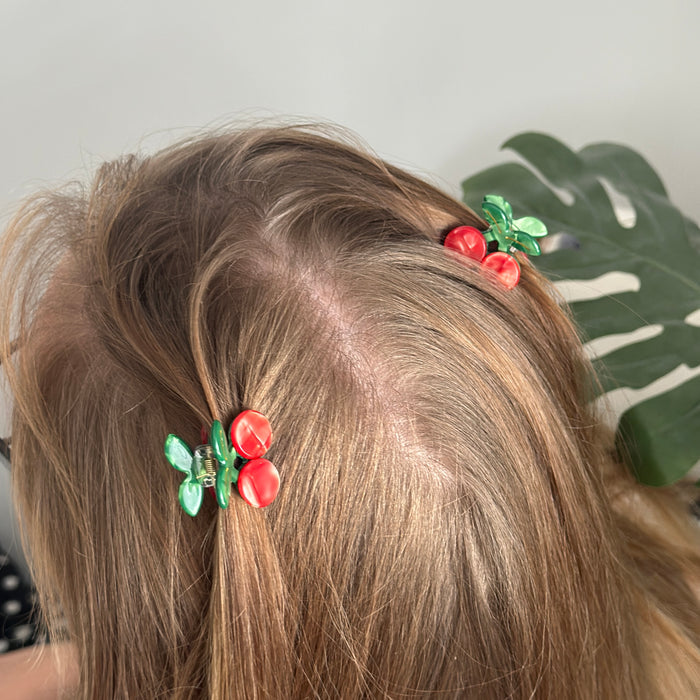 Light brown hair styled with Coconut Lane cherry-patterned mini claw clips; green plant in background.