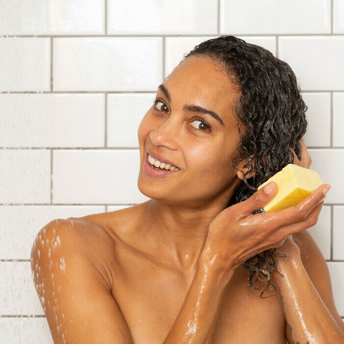Woman in shower with soap bar, white tiled wall background.