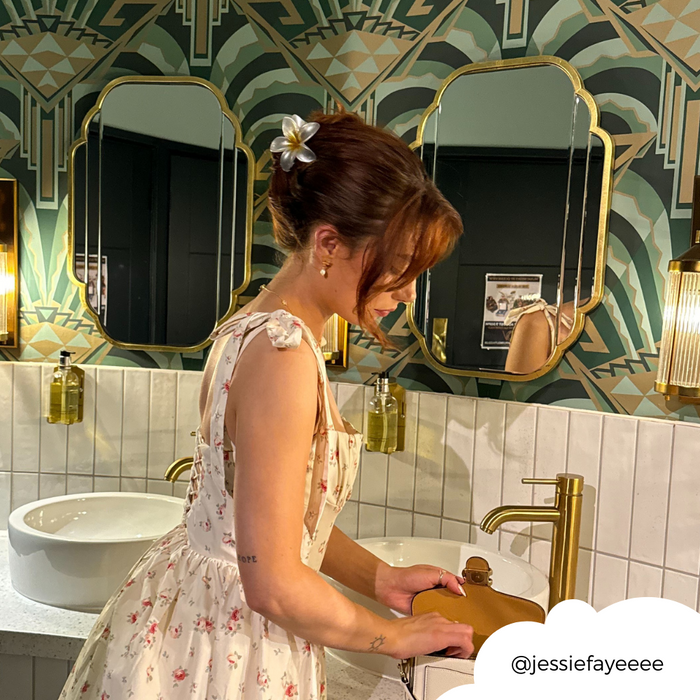 Woman in a floral dress wears a white tropical flower claw clip at a bathroom sink with gold fixtures.