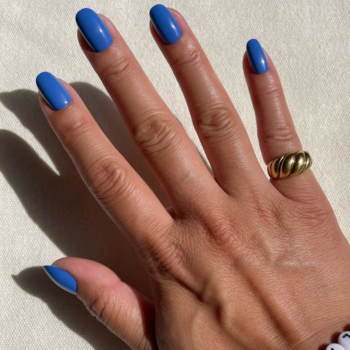Hand with Blue Canoe | Round | Short press-on nails in medium denim blue with glossy finish on natural skin tone against light fabric background