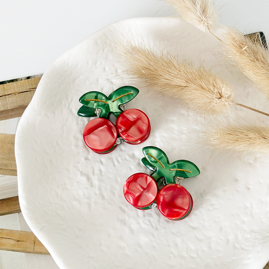 Cherry earrings and red cherry-shaped mini claw clips displayed on a white ceramic plate beside dried beige grass.