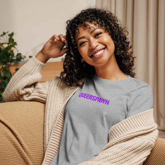 A smiling woman with curly hair sits on a beige couch wearing a light gray t-shirt with 'QUEERSPAWN' printed in purple across the chest, layered with a cream knit cardigan.