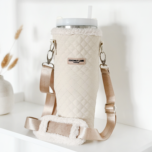 Quilted cream tumbler carry case with shearling trim and strap, displayed on a white shelf.